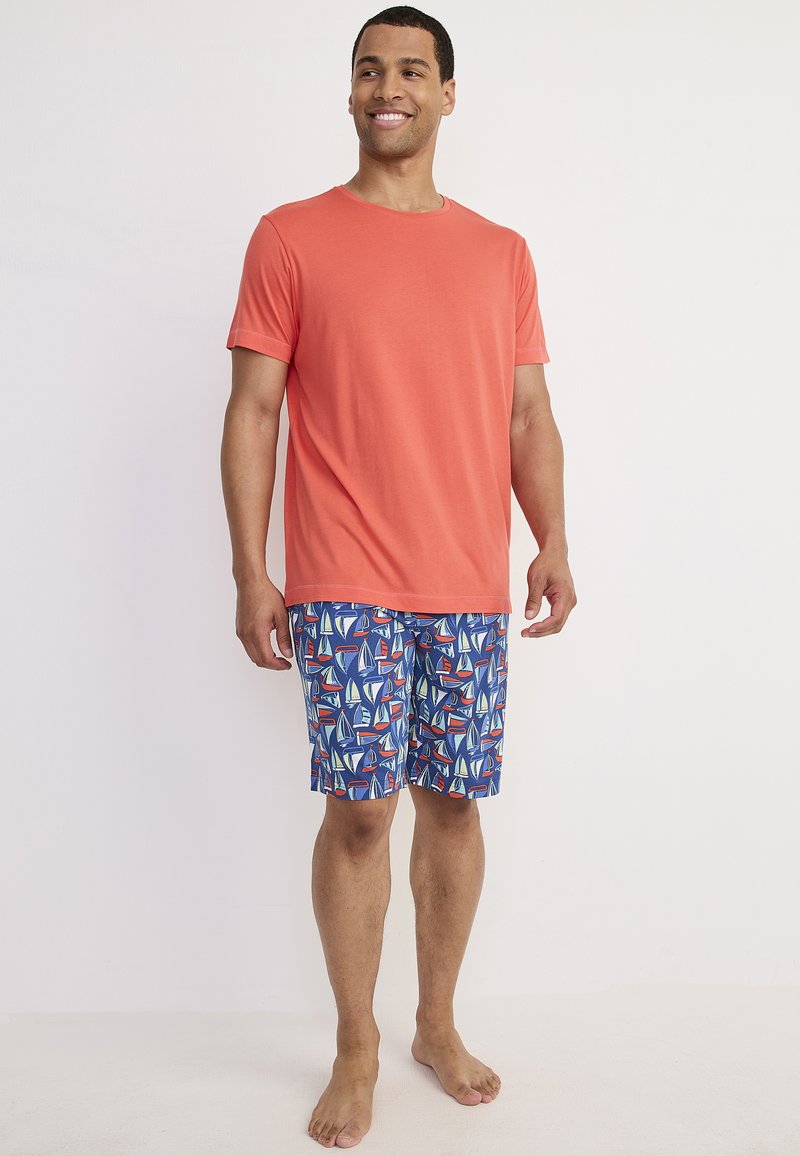 Korallfärgad t-shirt med rund halsringning, kombinerad med blå shorts med färgglada segelbåtsmönster. Modellen står barfota mot en enfärgad bakgrund.