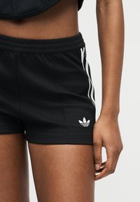 Άτομο που φοράει μαύρα σορτς Adidas με λευκές ρίγες και λογότυπο στο πλάι, εμφανίζοντας τη μέση και το δεξί χέρι με φόντο απλό.