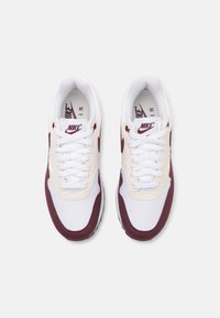 Nike Air tenisice u bijeloj boji s tamnocrvenim naglascima, sa gornjim dijelom od mreže, vezanjem na vezice i podstavljenim đonom. Tekstuirani bočni paneli.