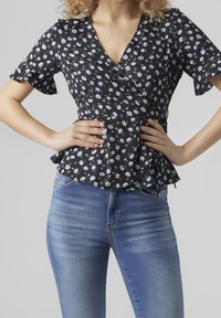 Top avvolgente nero con maniche corte arricciate, caratterizzato da piccoli motivi floreali blu e bianchi, abbinato a jeans azzurri chiari.