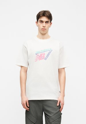 Fiatal férfi rövid barna hajjal, fehér New Balance futó pólót és sötét cargo nadrágot visel, egyszínű háttér előtt áll.