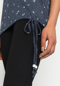 Blusa azul-marinho com estampado floral e bolinhas, apresentando uma bainha com cordão decorada com contas de madeira e azuis claras, combinada com calças pretas.