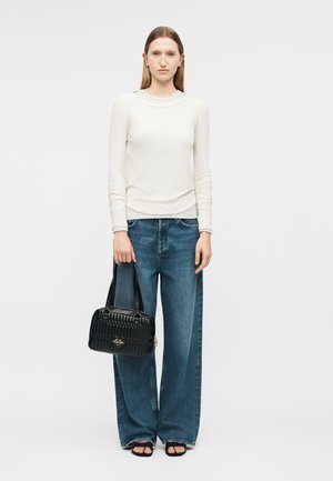 Moteris dėvi baltą ilgomis rankovėmis palaidinę, plačias mėlynas džinsines kelnes, juodus atvirus sandalų su atvirais pirštais, laiko juodą siuvinėtą rankinę su logotipu.