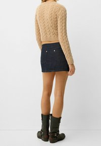 Bronzový pletený svetr s copánkovým vzorem; tmavě modrá džínová minisukně; zelené kotníkové boty s přezkami a mírným podpatkem; viditelné detaily zezadu.