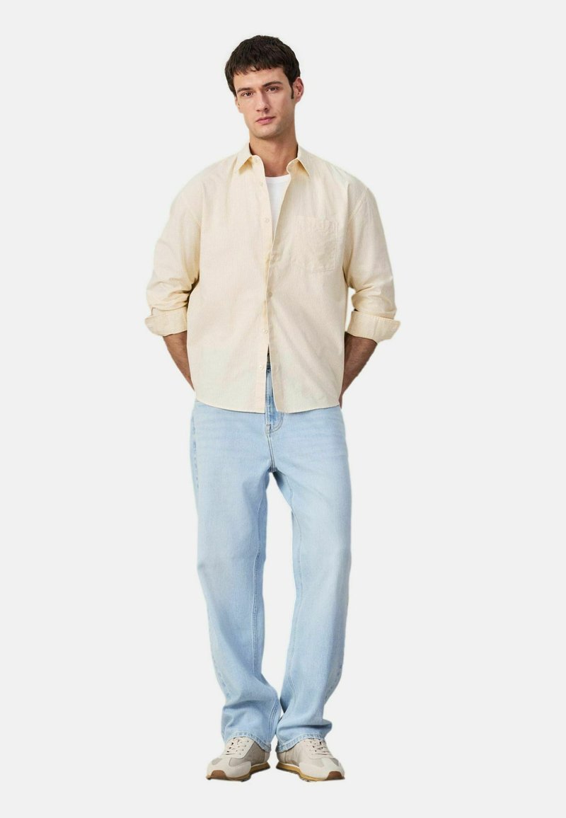 Jonge man die met de handen achter de rug staat, gekleed in een lichtbeige overhemd met knopen, een wit T-shirt eronder, lichtblauwe spijkerbroek en beige sneakers.