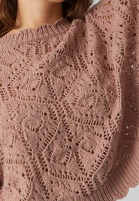 Frau trägt einen staubigen rosa Strickpullover mit detailliertem, offenem Rautenmuster und Blattmuster sowie einem gerippten, runden Ausschnitt.