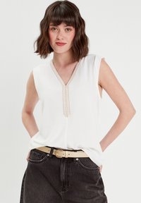 Haut sans manches blanc avec un col en V, orné de coutures décoratives le long de l'encolure ; porté avec un jean sombre taille haute et une ceinture beige.