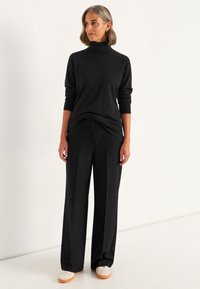 Frau, die auf einem weißen Hintergrund steht, wearing einen schwarzen Rollkragenpullover, schwarze weit geschnittene Hose, weiße Turnschuhe sowie goldene Ohrringe und ein Armband.