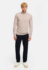 Ljusbeige zip-krage tröja med långa ärmar, kombinerad med svarta byxor och vita sneakers. Tyget verkar mjukt med en jämn textur.