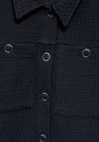 Camicetta nera con tessuto a trama, dotata di colletto, due tasche frontali e bottoni circolari con la scritta "CECIL."