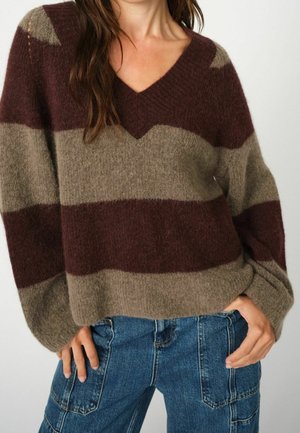 Mujer con suéter de rayas anchas en tonos marrón y topo, con cuello en V y tejido peludo, y jeans azules, con las manos en los bolsillos, sobre un fondo liso.