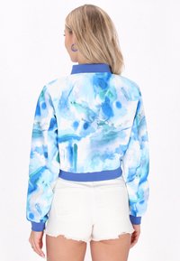 Giacca bomber cropped con un motivo astratto in acquerello blu e bianco, con polsini e colletto a coste blu, abbinata a pantaloni corti bianchi.