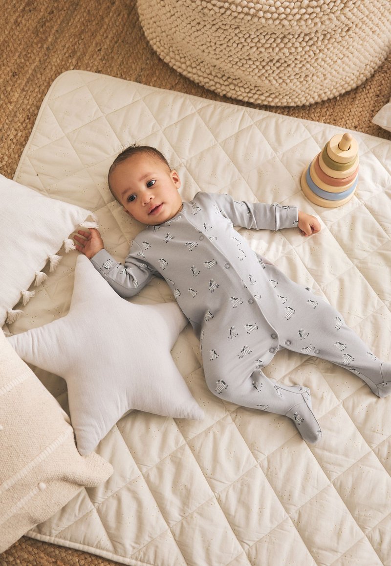 Baby in grey zebra-patterned onesie lying on quilted mat with star-shaped pillow and wooden stacking toy nearby.