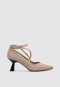 Zapatos de tacón de ante beige con punta afilada y tira en el tobillo. Presentan un diseño decorativo de trenzado cruzado y un tacón negro esculpido.