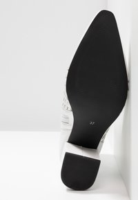 Bottine blanche avec semelle noire pointue et talon bloc, taille 37 indiquée sur la semelle, positionnée verticalement sur un fond blanc.