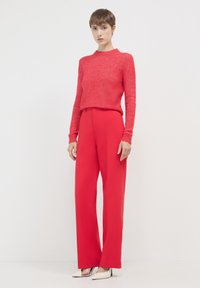 Roter, strukturierter Strickpullover und weite rote Hose, ergänzt durch spitze schwarz-weiße High Heels. Einfaches, klares Design mit moderner Silhouette.