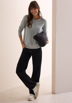 Lichtgrijze lange mouw top, donkerblauwe wijde broek en witte sneakers. Houdt een zwart gewatteerd item vast. Schone, minimale stijl tegen een neutrale achtergrond.