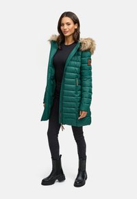 Abrigo acolchado verde con capucha forrada de pelo, cierre de cremallera, bolsillos laterales y textura acolchada. Lleva puesto con vaqueros ajustados negros y botas negras.