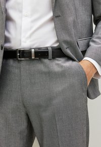 Szary garnitur o delikatnej fakturze, z białą koszulą pod spodem. Na talii widoczny czarny pasek z srebrną klamrą.