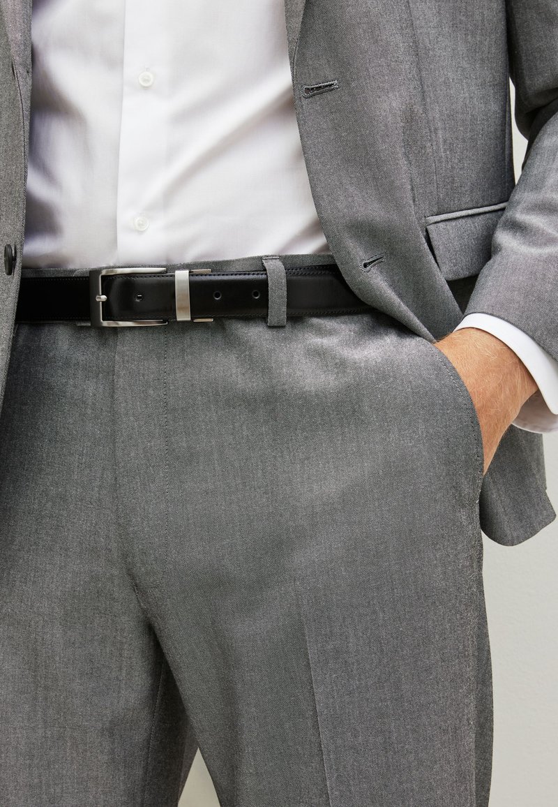 Szary garnitur o delikatnej fakturze, z białą koszulą pod spodem. Na talii widoczny czarny pasek z srebrną klamrą.
