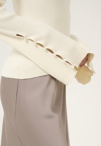 Crème ribbelsweater met parelaccenten langs de splitmouwen, gecombineerd met een gladde, taupe rok. Elegante en textuurrijke ontwerpelementen.