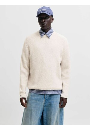 Mørkhudet person iført en blå kasket, cremefarvet sweater over en blåternet skjorte og løse lyseblå jeans, stående mod en ensfarvet baggrund.