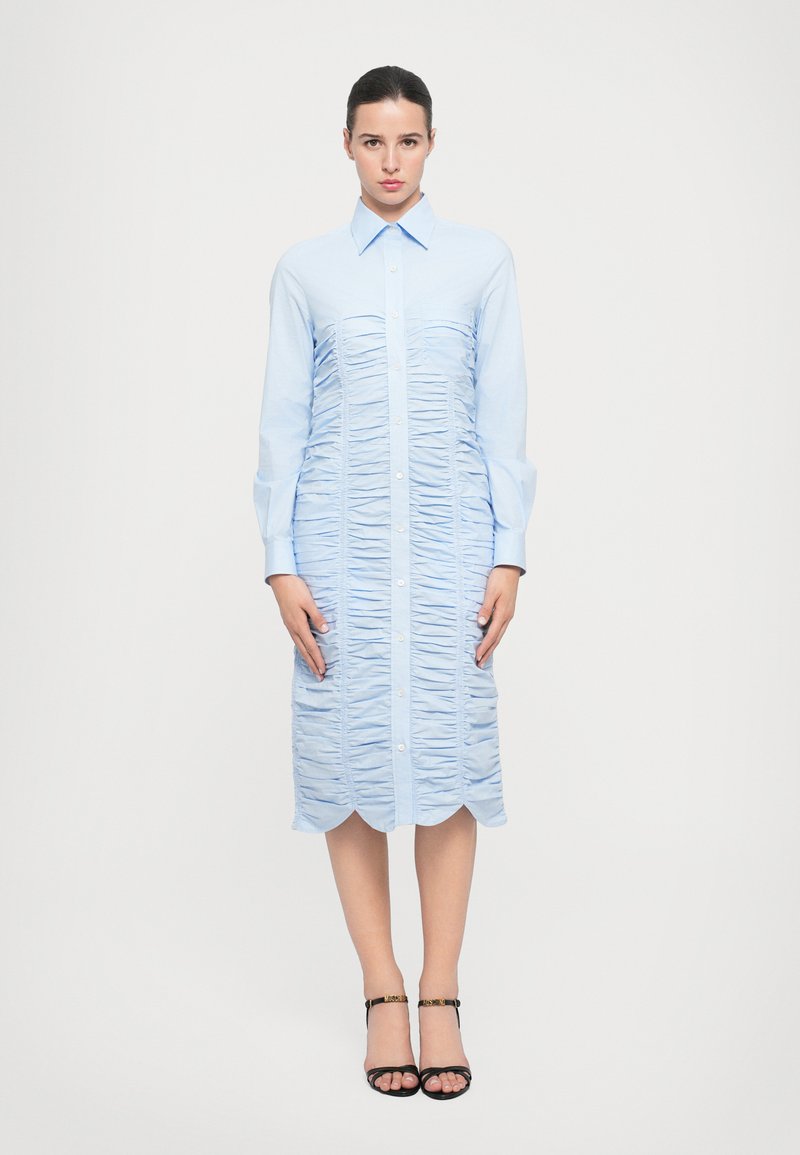 Rochie cămașă de culoare bleu deschis, cu un față cu nasturi, textură plisată, mâneci lungi și tiv ondulat. Purtați-o cu sandale negre cu toc.