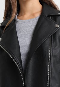 Giacca da moto in ecopelle nera con hardware argento, dotata di un ampio collo, zip diagonale e finitura testurizzata. Indossata sopra una maglietta grigia.