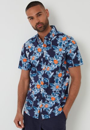 Mężczyzna w granatowej koszuli na krótki rękaw z dużym niebiesko-pomarańczowym kwiatowym wzorem, stojący na białym tle.