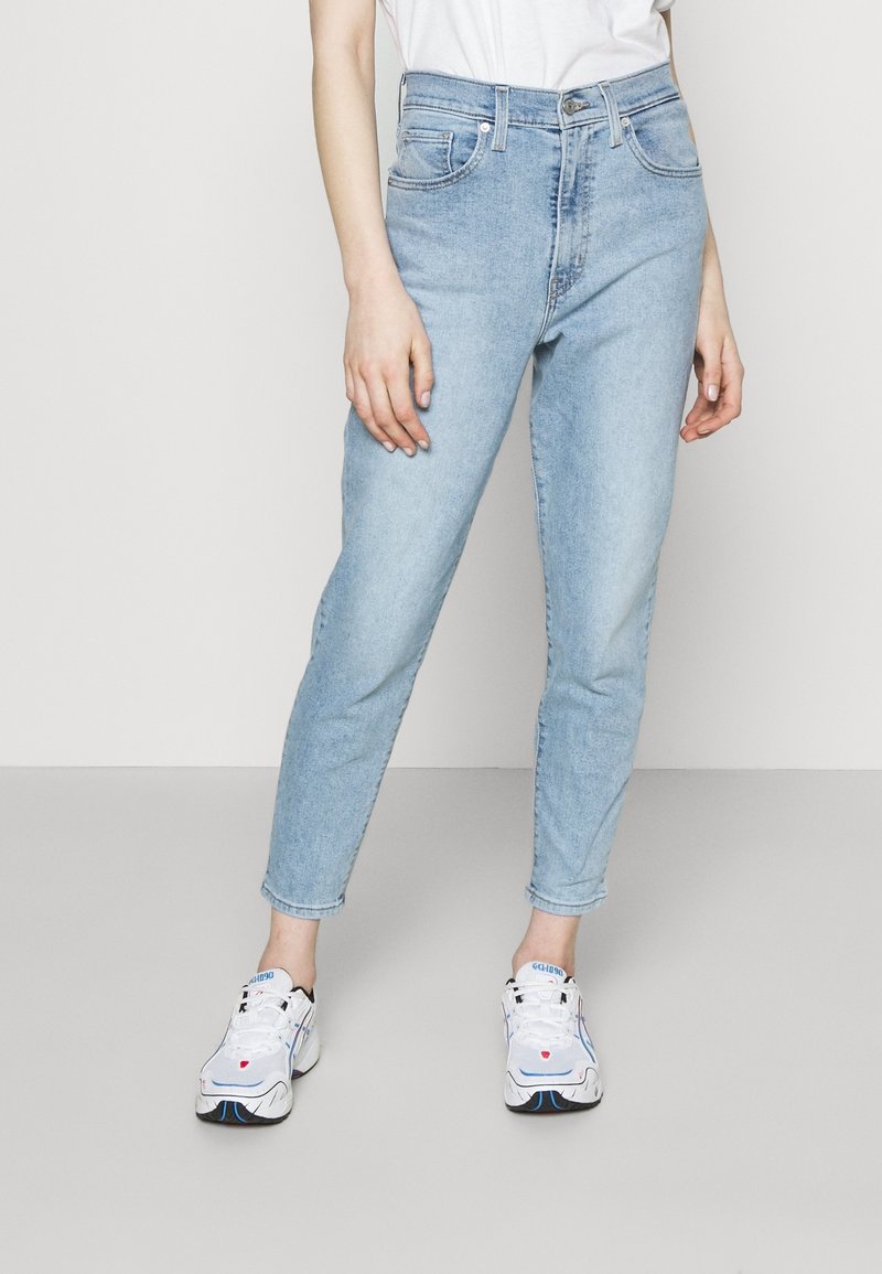 Person som bär ljusblå höga jeans, vita sportskor med blå och röda detaljer, och en vit topp, stående mot en enfärgad bakgrund.