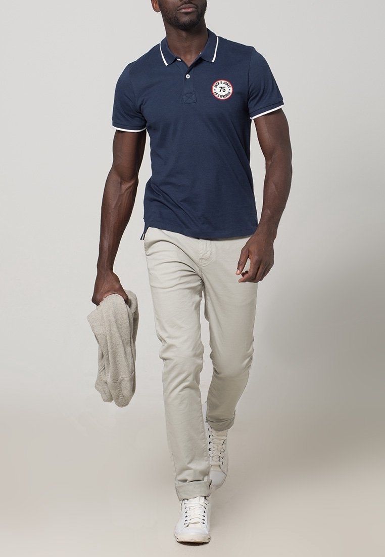 Homme marchant portant un polo bleu marine, un pantalon beige clair, des baskets blanches, tenant un pull beige dans sa main gauche.