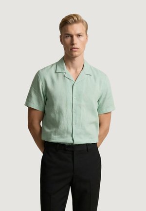 Uomo biondo con capelli corti che indossa una camicia verde chiaro a maniche corte con bottoni e pantaloni neri, in piedi con le mani dietro la schiena.