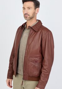 Braune Lederjacke mit Kragen, gerippten Bündchen und einer Fronttasche mit Knopf. Hergestellt aus glattem, strukturiertem Leder.