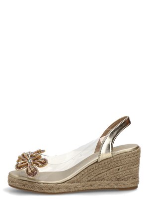 Sandalia de cuña dorada con tacón de cuerda trenzada, tiras transparentes y adorno floral decorativo con cuentas en la punta.