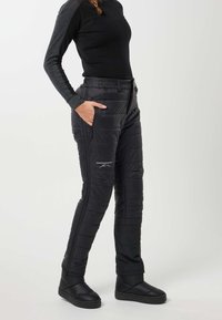 Henkilö, jolla on mustat tikatut ulkohousut vetoketjutaskuilla ja mustat slip-on-saappaat, seisoo yhdellä kädellä taskussa.