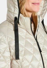 Giacca trapuntata beige chiaro con colletto alto, caratterizzata da una zip nera e cordini a contrasto neri con testo in rilievo lungo i lati.