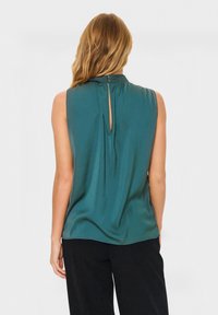 Bežė rankovių melsvai žalia bluzonė su aukštu kaklu, susirinkusia nugaros detale ir rakto skylės atidarymu, pagaminta iš lygios, plaukiančios medžiagos.