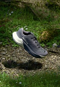 Chaussure de sport grise avec une tige texturée, des accents blancs et une semelle robuste, présentée sur un fond de gazon naturel et de pierres.