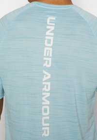 Camisa atlética de color azul claro con un patrón texturizado, con 'UNDER ARMOUR' impreso verticalmente en blanco en la parte de atrás.