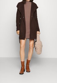 Lang brun cardigan med draperet krave, båret over en lysebrun kjole. Modellen holder en fuzzy beige taske og har glossy brune ankelstøvler på.