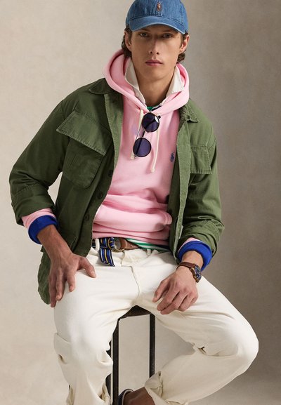 Jaunas vyras sėdi ant kėdės, dėvi mėlynę kepuraitę, žalią striukę ant rožinės džemperio su gobtuvu, baltas kelnes, saulės akinius kabinančius ant džemperio ir laikrodį kairėje riešo pusėje.