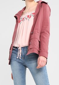 Donna che indossa jeans blu sbiaditi, una maglietta floreale rosa off-shoulder con cordino e una giacca rosa polvere con zip e tasche frontali.