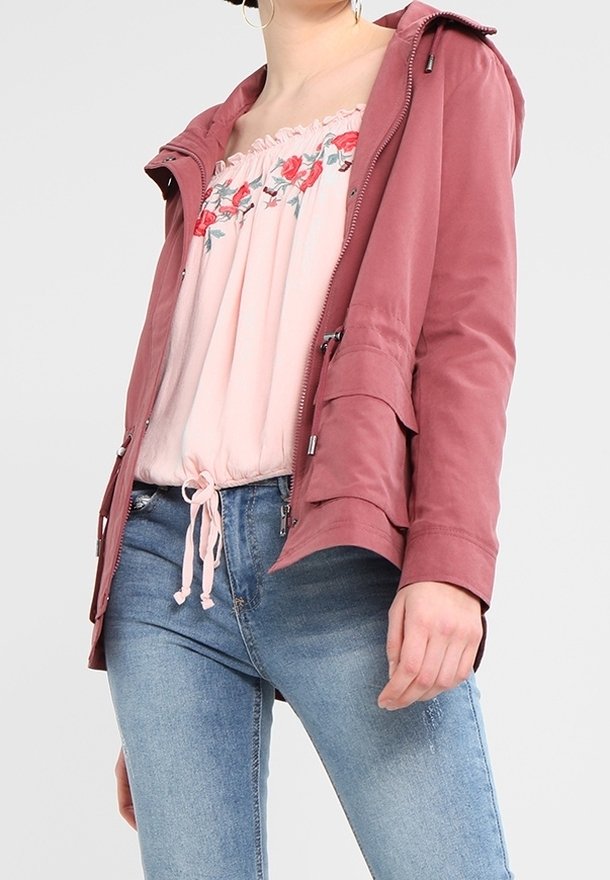 Donna che indossa jeans blu sbiaditi, una maglietta floreale rosa off-shoulder con cordino e una giacca rosa polvere con zip e tasche frontali.