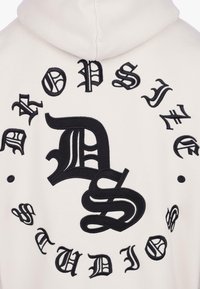 Biela mikina s kapucňou s veľkými čiernymi vyšívanými gotickými písmenami tvoriacimi "Dropsy Studio" v kruhu okolo prekrývajúcich sa iniciálok "D" a "S".