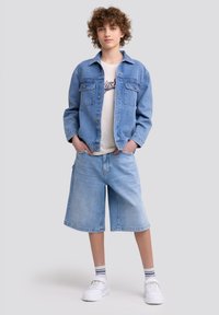 Giacca di jeans azzurro chiaro con due tasche sul petto, abbinata a pantaloni shorts di jeans azzurro chiaro. Sneakers bianche e una T-shirt bianca con testo grafico.