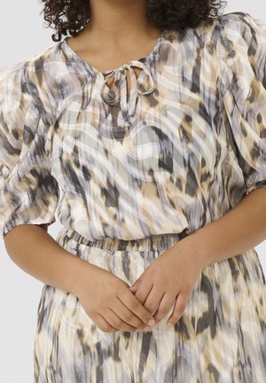 Femme portant une robe beige, grise et blanche à motifs, manches courtes bouffantes et col noué, les mains doucement jointes à la taille.