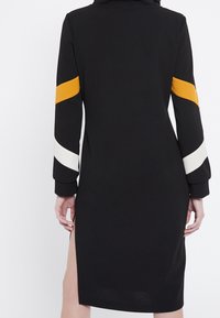 Svart klänning med långa ärmar, prydd med gula och vita ränder på ärmarna, en sidosprund och ett mjukt texturerat material.