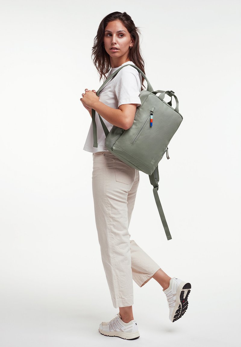 Zaino in tessuto verde con spalline regolabili, maniglie superiori, una tasca anteriore con zip e dettagli hardware colorati. Il modello posa in abbigliamento casual.