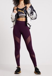Top sportivo corto nero e oro, giacca imbottita bianca e nera, leggings viola con pannelli in rete e motivi diagonali, scarpe da atletica nere.
