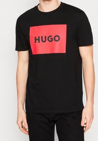 Sort kortærmet T-shirt med et iøjnefaldende rødt rektangulært grafisk design med ordet "HUGO" i sort, lavet af blødt bomuldsstof.